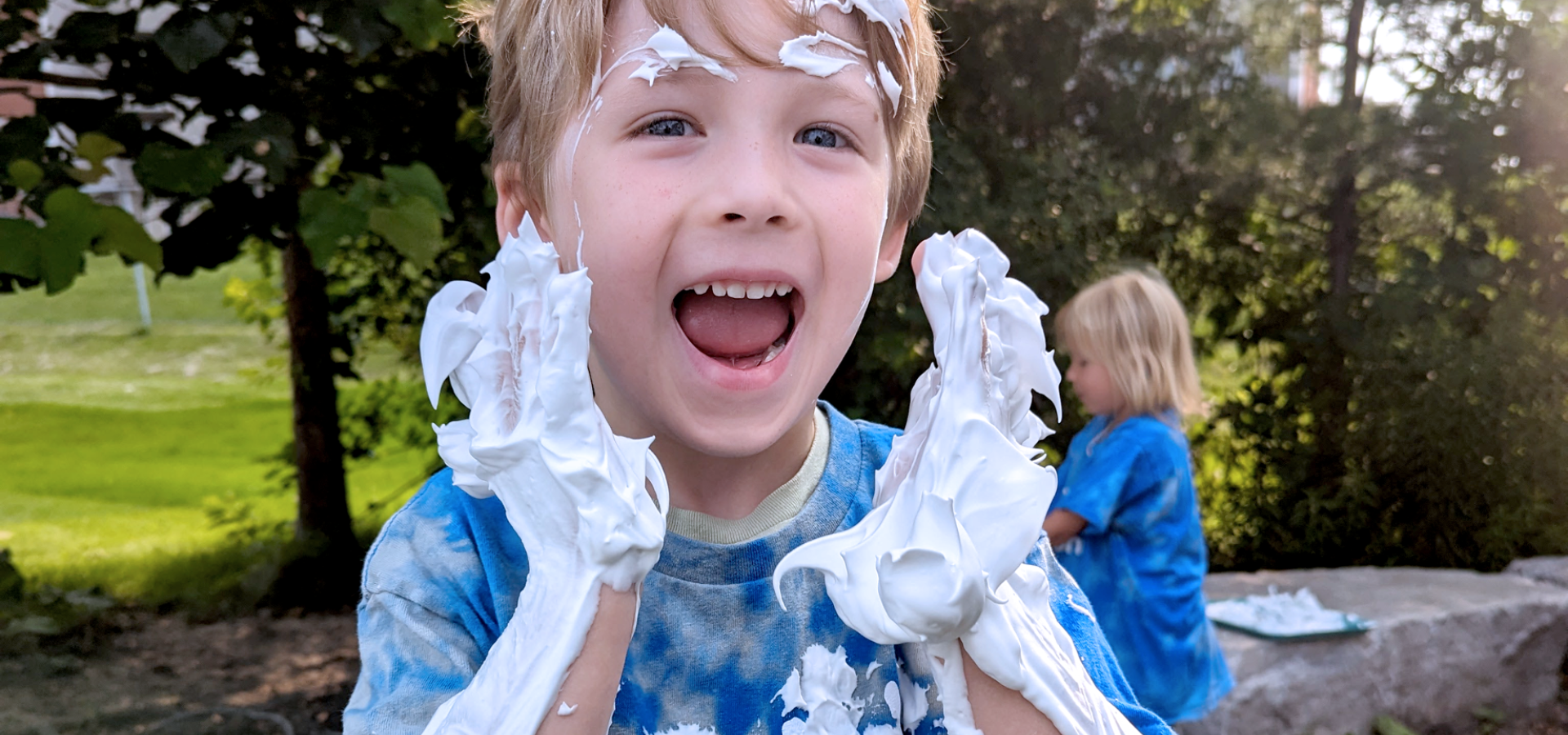 young boy with blonde hair in a royal blue london children's connection shirt covered in shaving cream as part of sensory activity