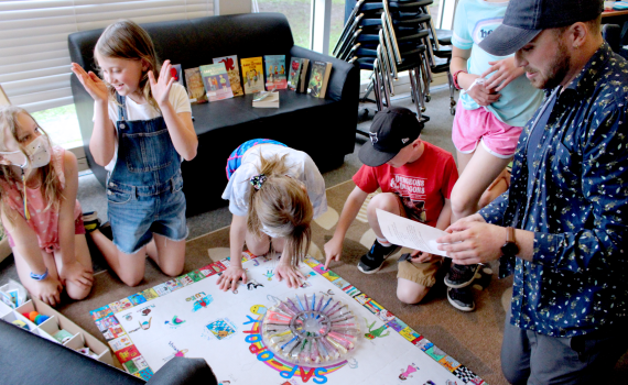 young male educator and group of school age children kneeling beside a homemade board game they created