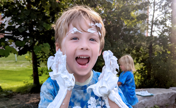 young boy with blonde hair in a royal blue london children's connection shirt covered in shaving cream as part of sensory activity