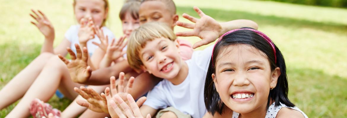 five multi-cultural school age children sit on grass and wave at camera