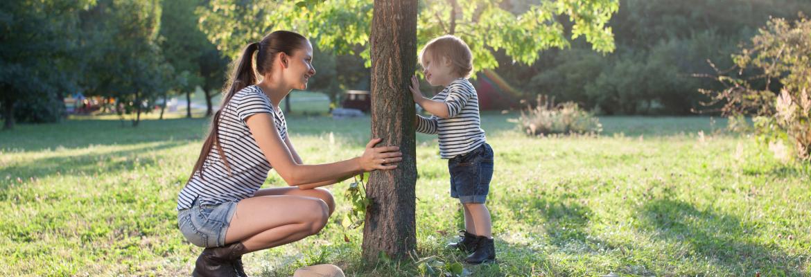 child and educator smiling across at each other while both holding on to base of a tree